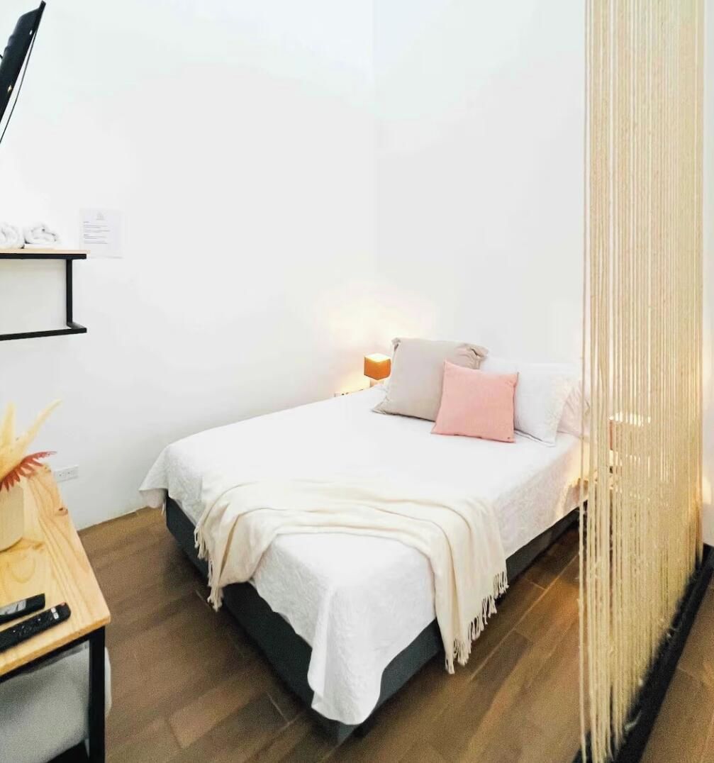 Un dormitorio con cama, mesita de noche, lámpara y cortina.