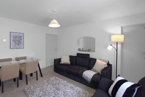 A modern living room with a dark sofa, a dining table with chairs, a large mirror, and a floor lamp. - Stylish 3BR Apartment | Clapham Junction | Parking (London)