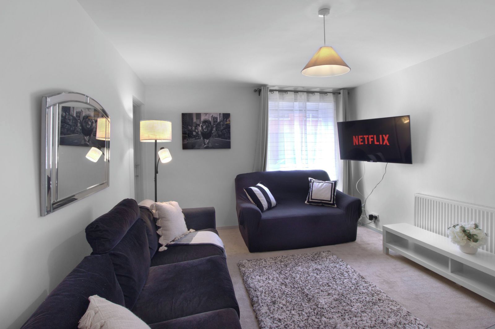 A modern living room with a dark sofa, a smaller armchair, a floor lamp, a flat-screen TV, and a large mirror on the wall.