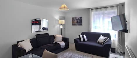 A modern living room with two dark sofas, a glass coffee table, a lamp, a flat-screen TV, and a window with curtains.