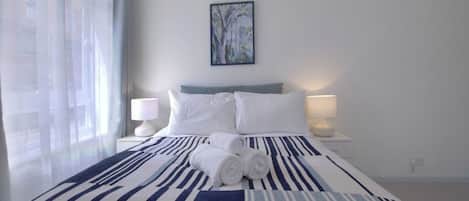 A hotel room with a bed, two bedside lamps, a painting on the wall, and a window with curtains.