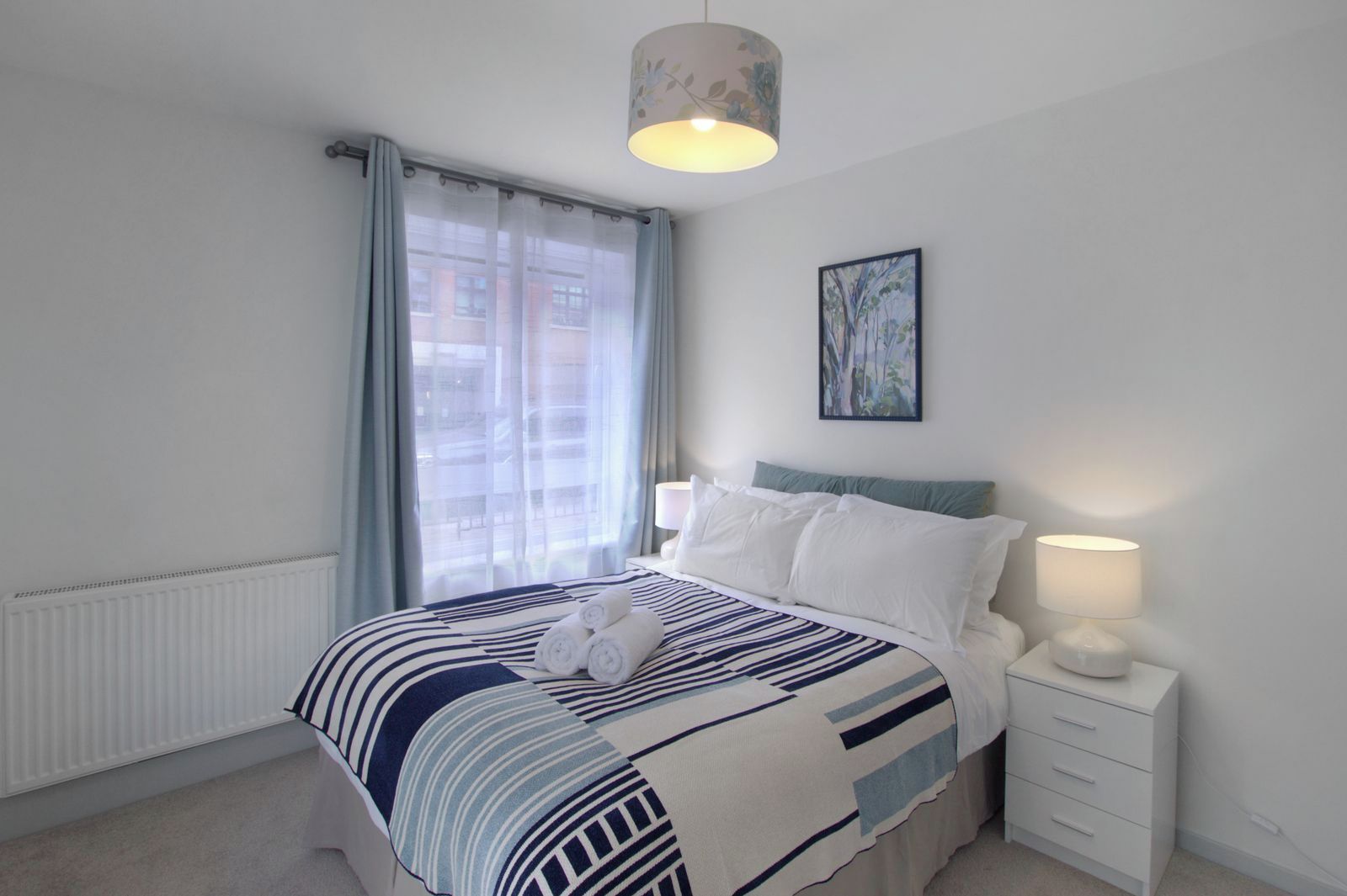 A bedroom with a bed, a nightstand, a lamp, a window with curtains, and a painting on the wall.