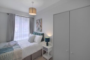 Ein Schlafzimmer mit einem Bett, einem Nachttisch mit Lampe, einem Kleiderschrank und einem Fenster mit Vorhängen.