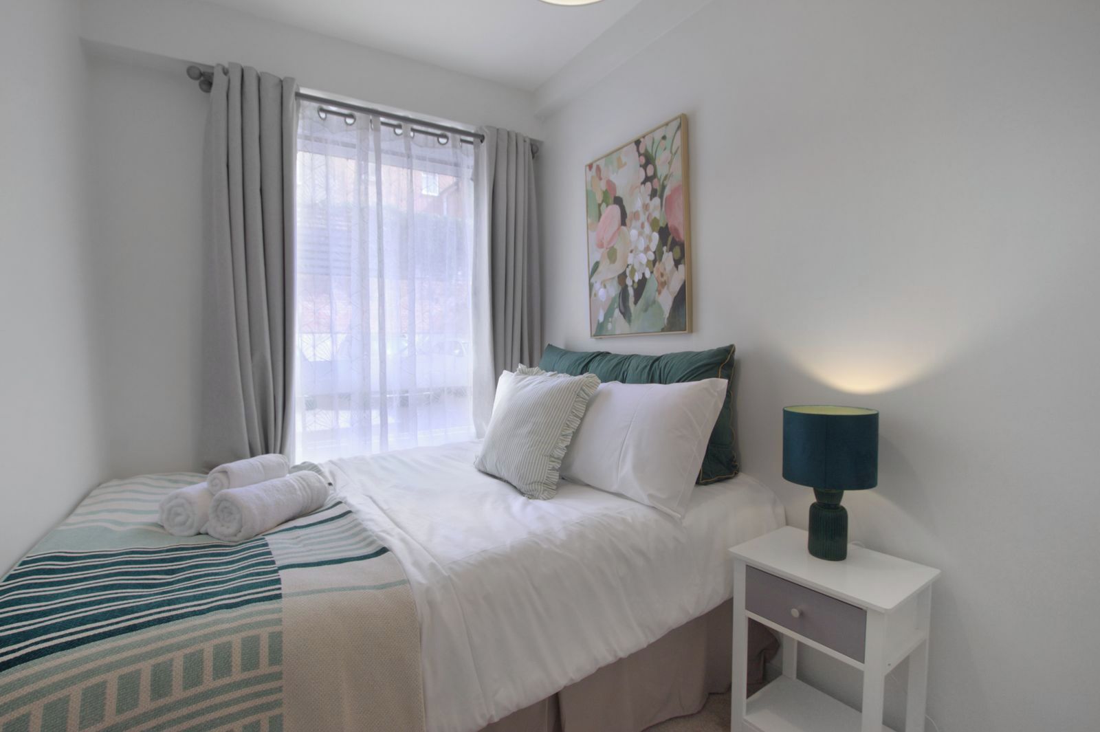 A hotel room with a bed, a nightstand with a lamp, a window with curtains, and a painting on the wall.
