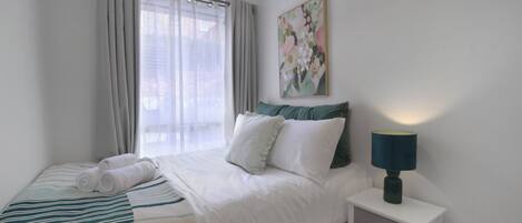 A hotel room with a bed, a nightstand with a lamp, a window with curtains, and a painting on the wall.