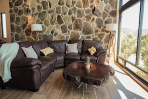 Una sala de estar moderna con una pared de piedra, sofás de cuero, una mesa de centro redonda y vistas a través de un ventanal grande.
