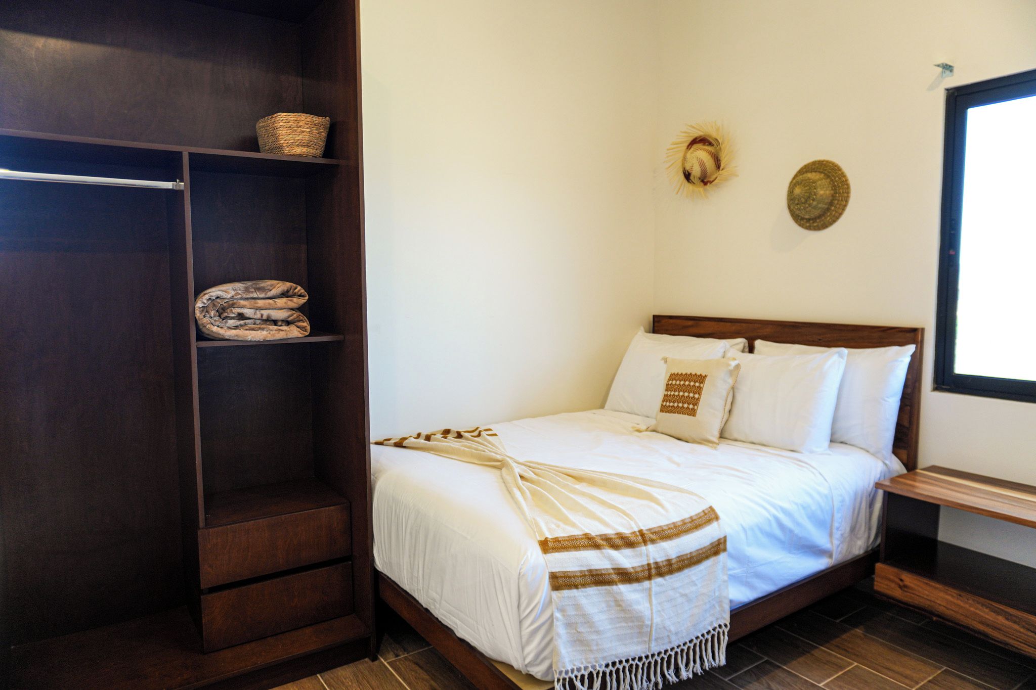 Uma cama bem arrumada com manta branca e marrom, cabeceira de madeira e uma prateleira com manta dobrada e um cesto.