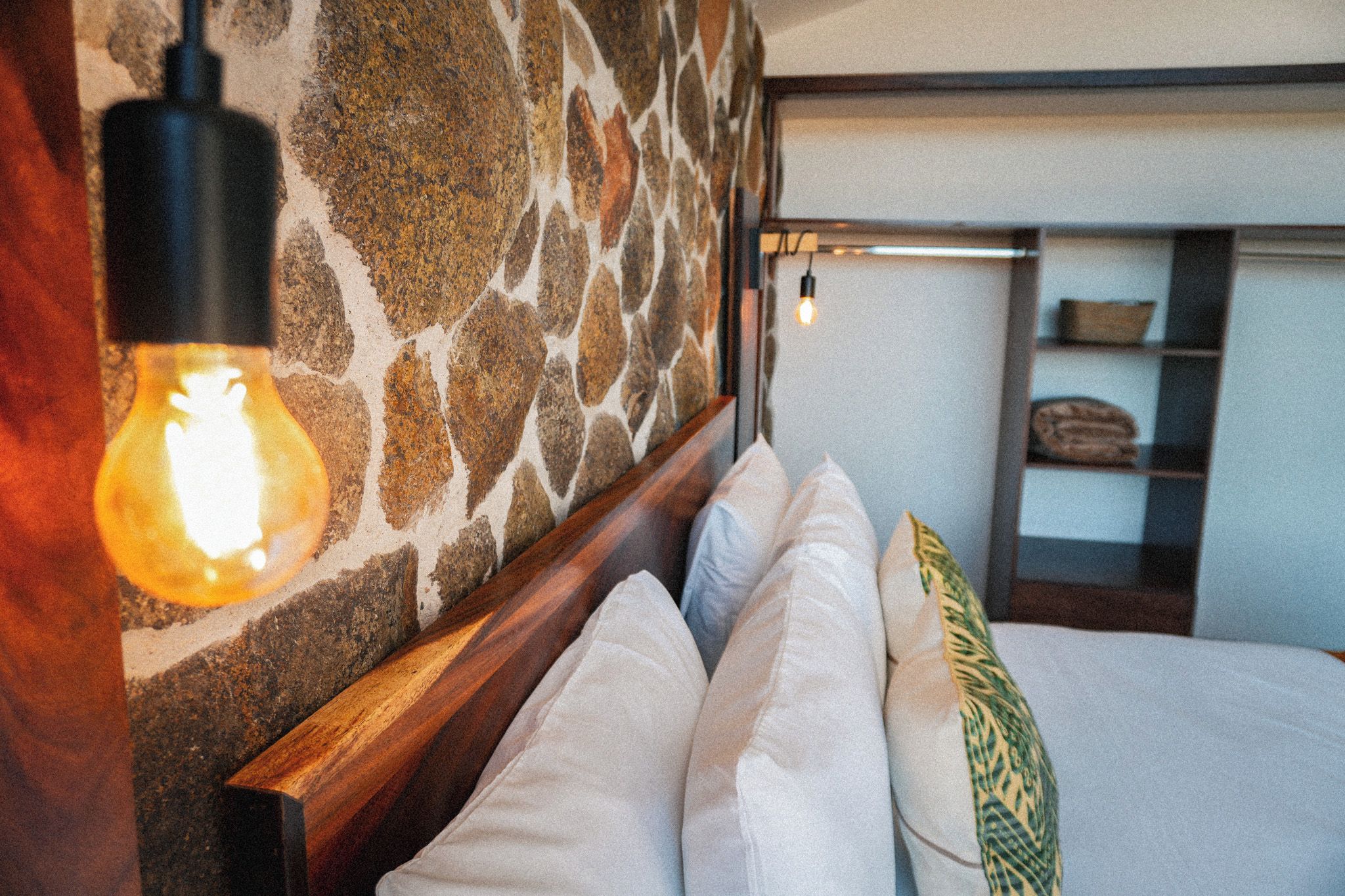 A bed with white and patterned pillows, a stone wall, and a hanging light fixture.