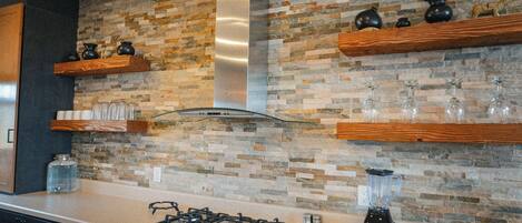 A kitchen with a gas stove, a blender, and a stone backsplash.