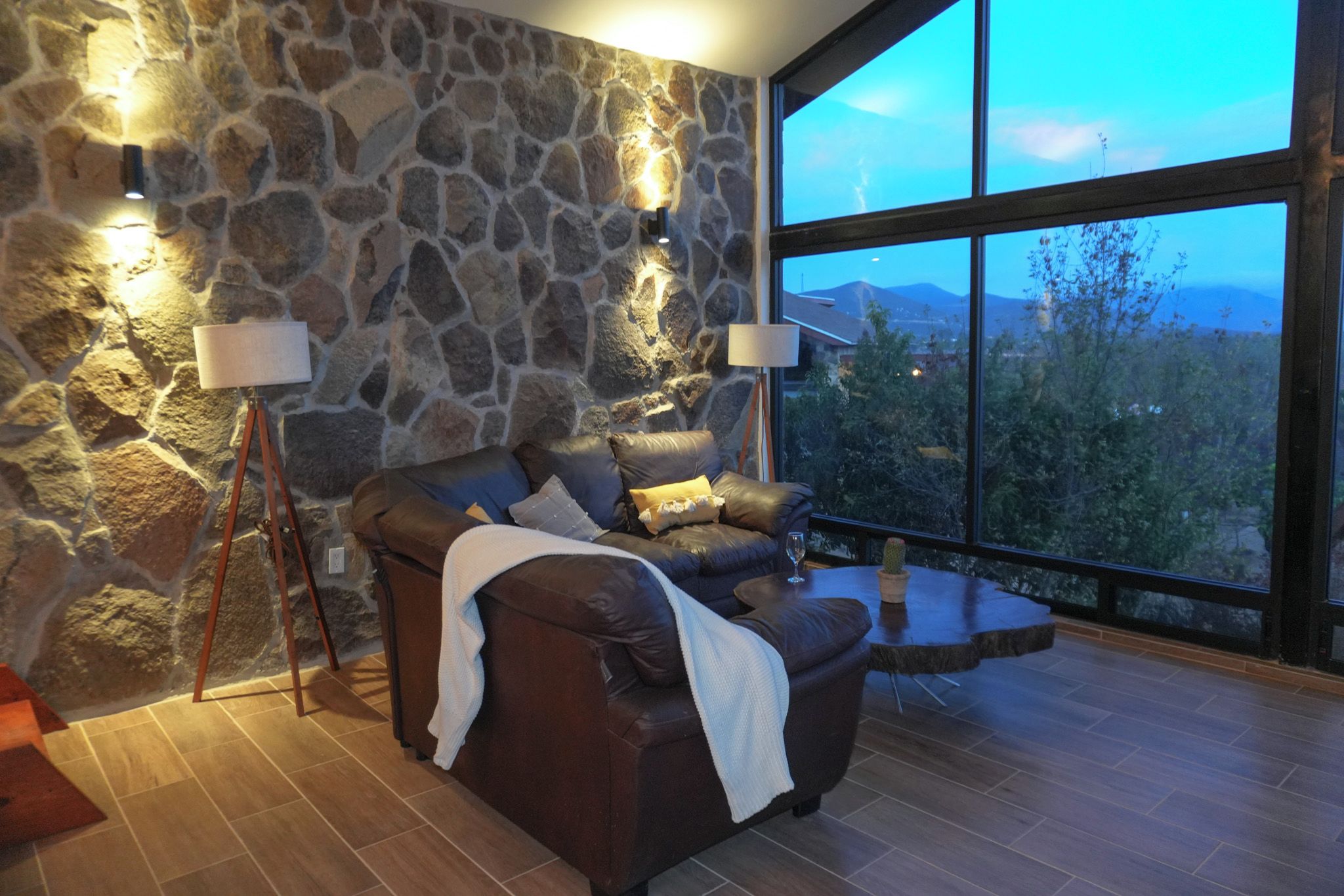 A stone-walled room with a leather sofa, two floor lamps, and a large window overlooking a mountainous landscape.