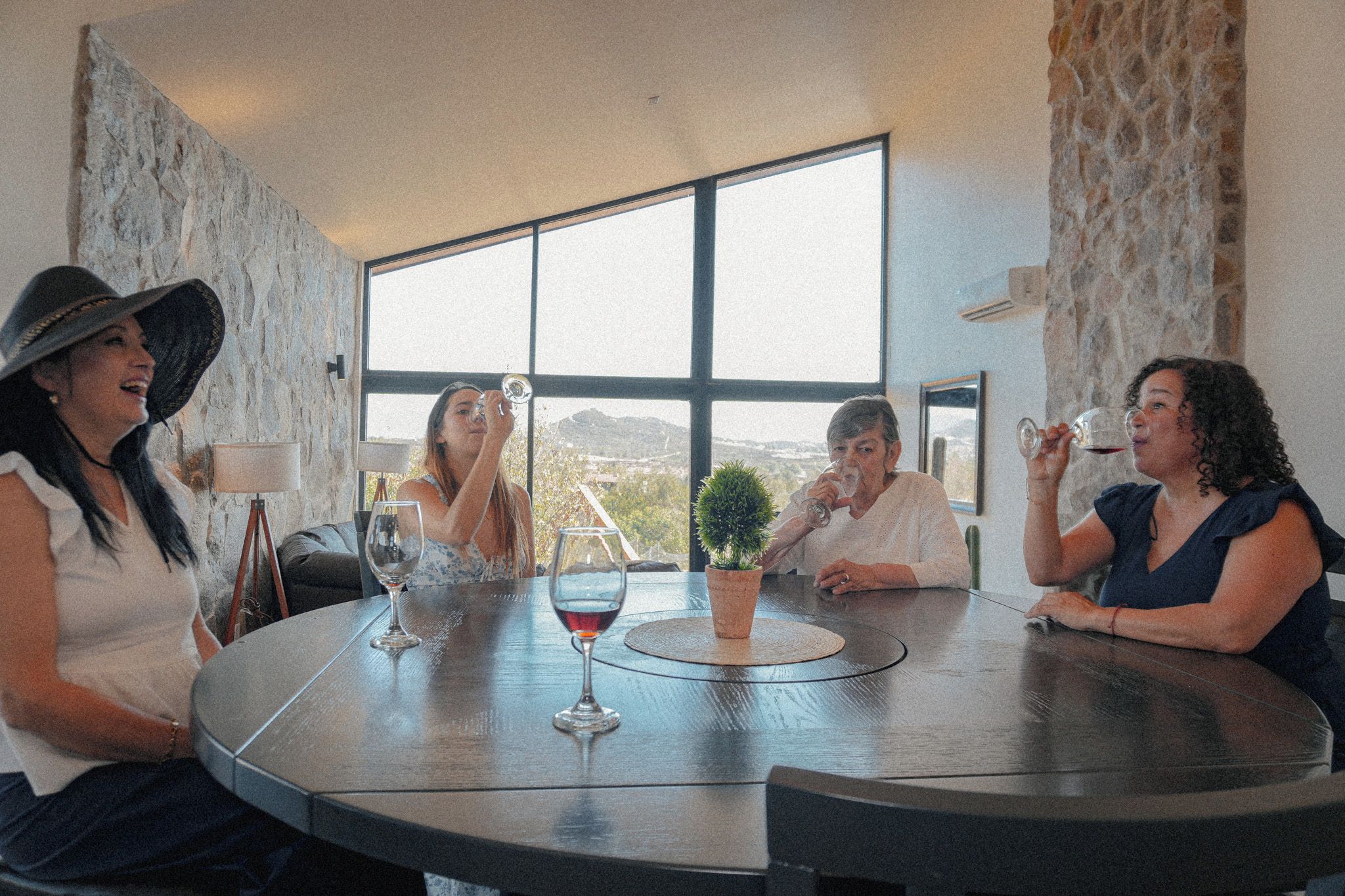 A group of people seated around a round table with wine glasses, in a room with a large window and stone walls.
