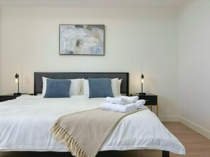 A neatly made bed with a beige throw blanket, two bedside lamps, and a modern abstract painting on the wall. - Panoramic Ocean Views – 4-Bedroom North Nanaimo Suite with Private Balcony (Nanaimo)