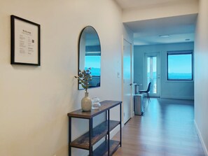 A hallway with a framed welcome sign, a vase with flowers on a console table, and a view of a room with a chair and a window. - Panoramic Ocean Views – 4-Bedroom North Nanaimo Suite with Private Balcony (Nanaimo)