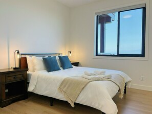 A neatly made bed with white bedding and blue pillows, a bedside table with a lamp, and a large window with a view. - Panoramic Ocean Views – 4-Bedroom North Nanaimo Suite with Private Balcony (Nanaimo)