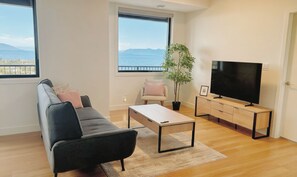 Un salon moderne comprenant un canapé, une table basse, un téléviseur et une grande fenêtre donnant sur la mer et les montagnes.