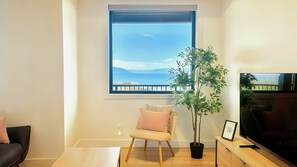 A living room with a large window, a potted plant, a chair, a coffee table, and a TV. - Panoramic Ocean Views – 4-Bedroom North Nanaimo Suite with Private Balcony (Nanaimo)
