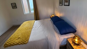Un dormitorio con cama, lámpara de noche, ventana con cortinas y dos cuadros en la pared.