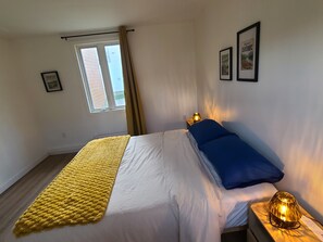 Een slaapkamer met een bed, een bedlamp, een raam met gordijnen en twee ingelijste foto's aan de muur.