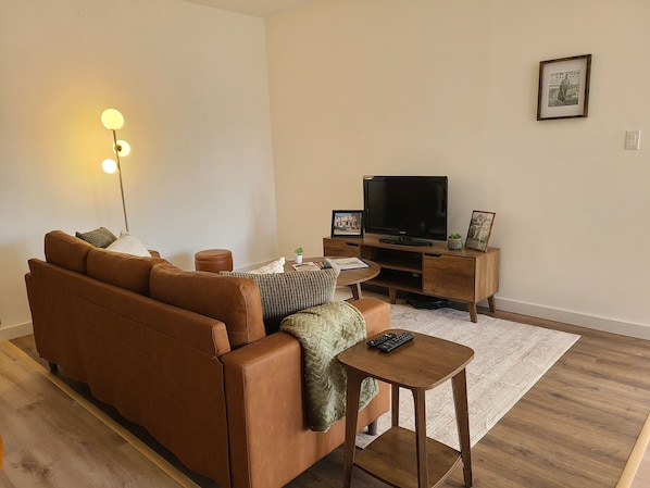 Een woonkamer met een bruine leren bank, een flatscreen televisie op een houten standaard, een klein houten bijzettafeltje en een vloerlamp.