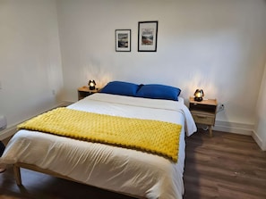 A bedroom with a bed, two bedside tables, a yellow blanket, and two framed pictures on the wall.