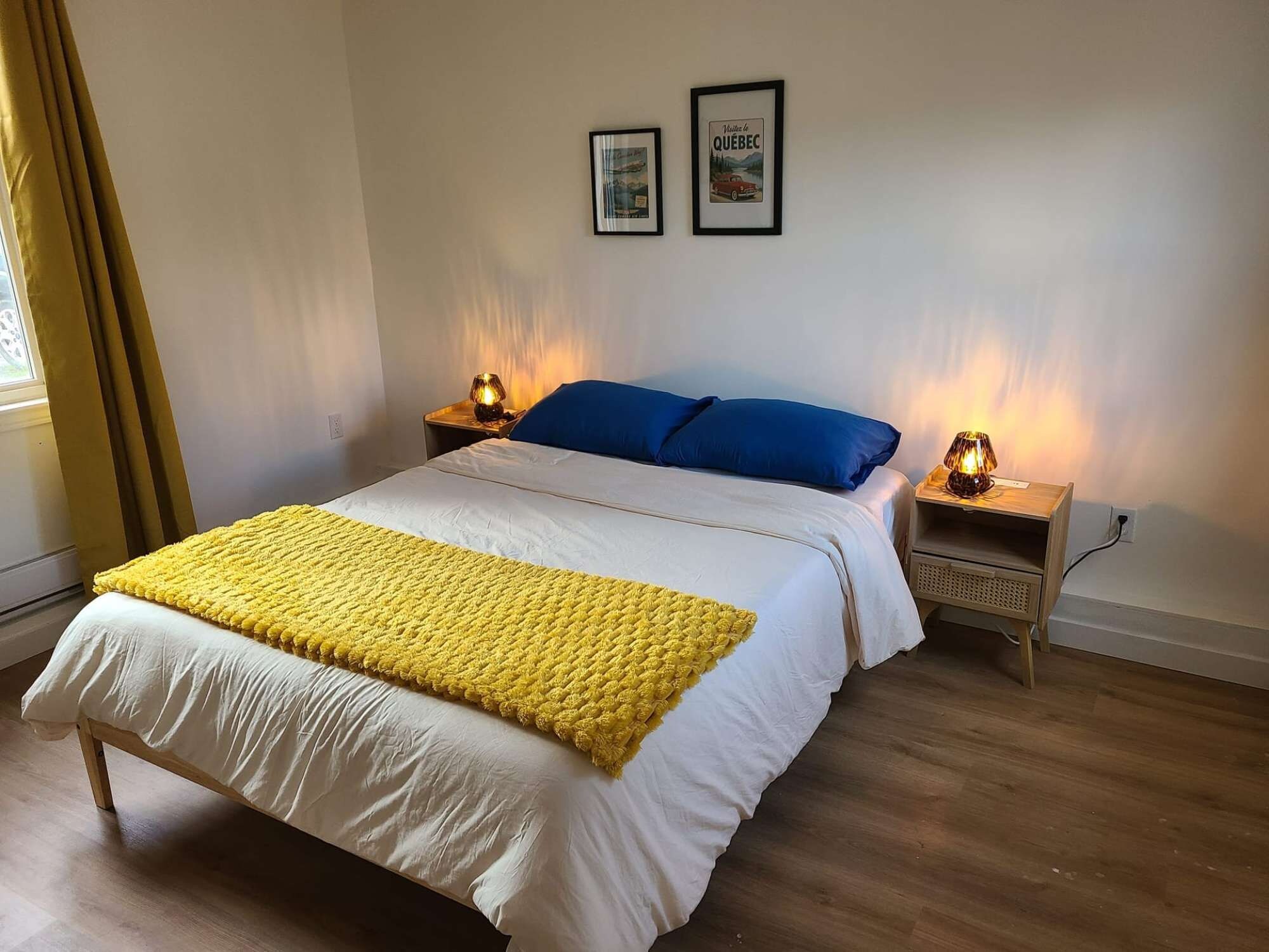 Une chambre à coucher avec un lit, deux lampes de chevet, un plaid jaune et deux tableaux encadrés accrochés au mur.