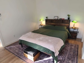 A bedroom with a bed, two bedside tables, a nightstand, a small wooden chest, and a patterned rug.