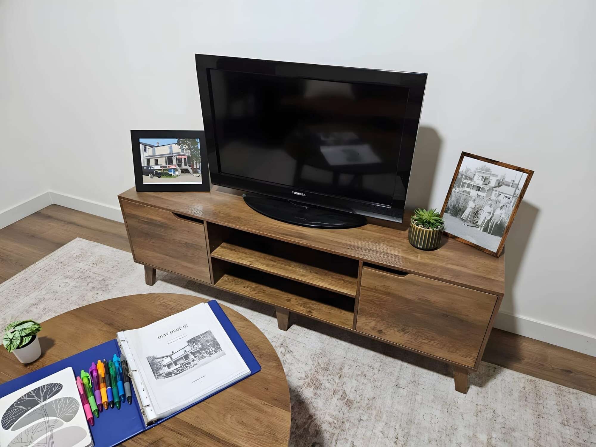 A flat-screen TV on a wooden entertainment center with two framed pictures and a book with pens on a round coffee table.