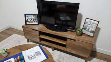 A flat-screen TV on a wooden entertainment center with two framed pictures and a book with pens on a round coffee table.