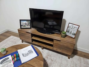 A flat-screen TV on a wooden entertainment center with two framed pictures and a book with pens on a round coffee table.
