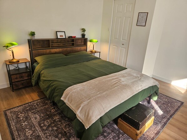 A bedroom with a wooden headboard, green bedding, two bedside lamps, a small shelf with decorative items, and a patterned rug.