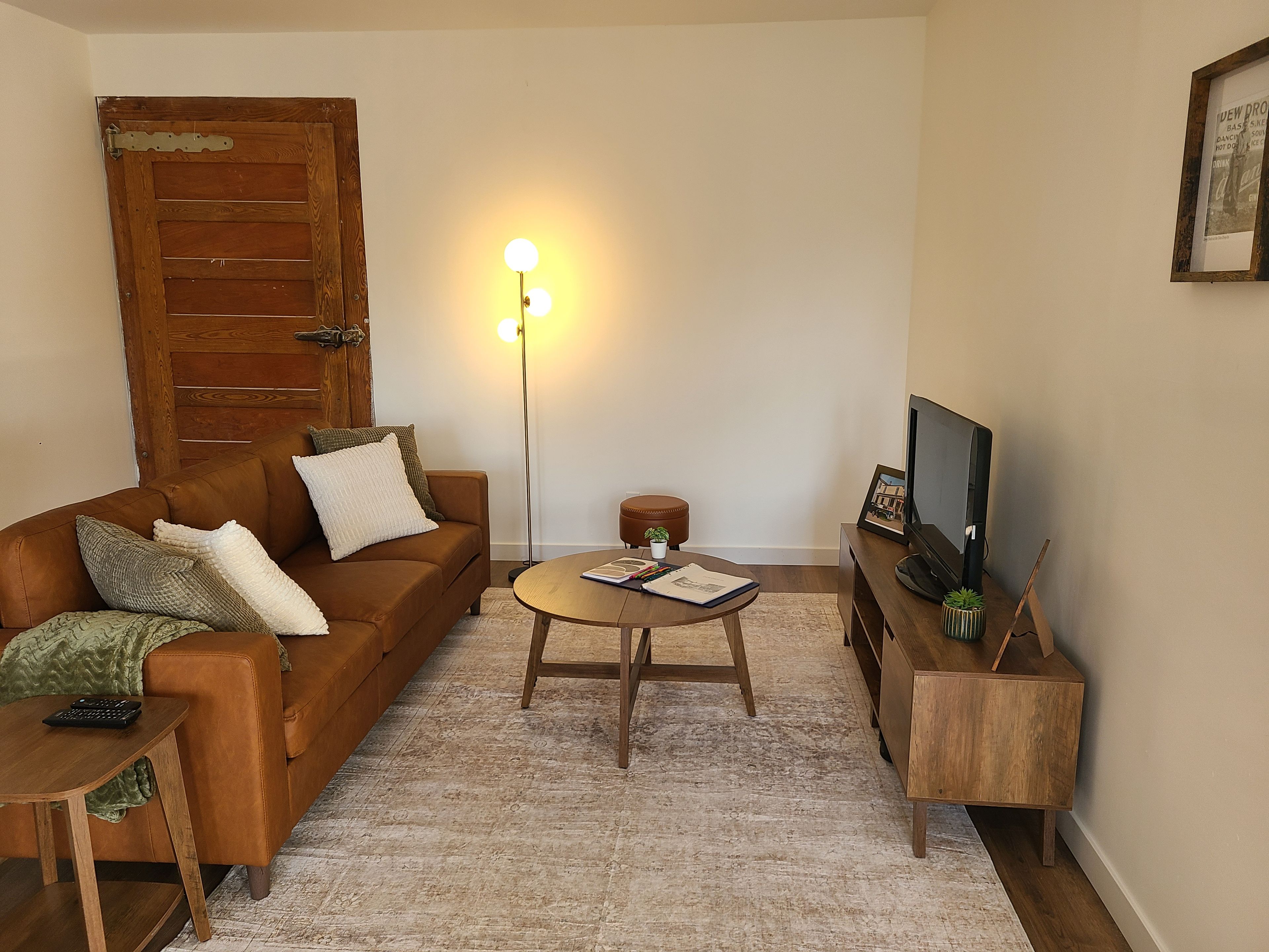 Uma sala de estar com um sofá de couro marrom, uma mesa de centro redonda de madeira, uma televisão sobre um suporte de madeira e um abajur com três lâmpadas.
