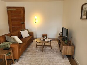 Una sala de estar con un sofá de cuero marrón, una mesa de centro redonda de madera, un televisor sobre un mueble de madera y una lámpara de pie con tres luces.