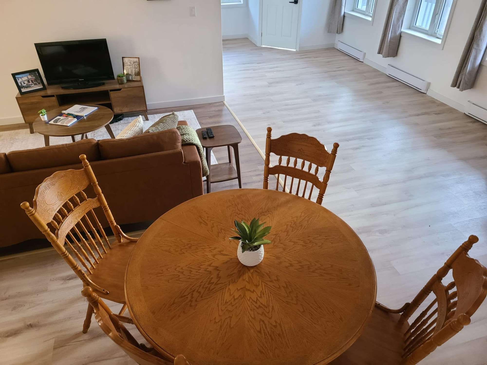 Una sala de estar con un sofá marrón, una mesa de comedor de madera con sillas, un televisor y una pequeña planta en maceta.