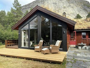 Eine moderne Hütte mit einer großen Glasfront, einer Holzterrasse, Korbmöbeln und einem Kiesweg.