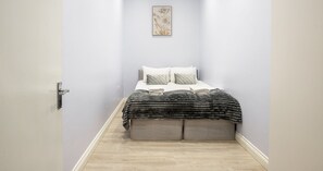 Ein schmaler Flur führt zu einem Bett mit einer grau-schwarz gestreiften Decke, einem grauen Kopfteil und zwei Kissen.