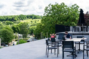 Een terras buiten met tafels en stoelen, met uitzicht op een groen landschap met bomen en heuvels.