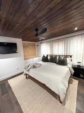 A bedroom with a wooden ceiling, a bed with white bedding, a ceiling fan, and a window with curtains.