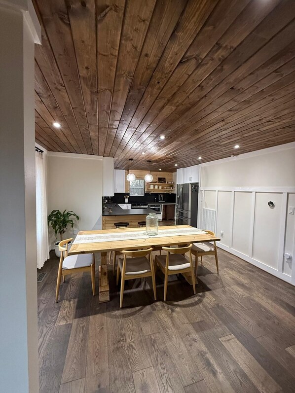 A modern kitchen with wooden ceiling, a dining area with a wooden table and chairs, and a view into the living space.