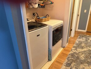 A compact laundry area with a washing machine and dryer, a basket, and a shelf with various items. - 3-bedroom stilt home designed for hunters, fisherman, scallopers & nature lovers (Crystal River)
