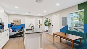 A modern kitchen with a central island, a dining area with a table and bench, and a large window with blinds. - The Reset Estate by Avantstay Book for ACL Sleeps 22 Pool + Hot Tub + Sauna Casita 2 Acres (Austin)