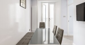 A modern dining area with a glass table and cushioned chairs, white walls, and a door leading to a balcony. - South Kensington Base Near Chelsea (London)