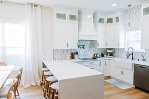 A modern kitchen with white cabinetry, a central island, and a dining area with wicker chairs. - The Cove & Beach Nook | Sleeps 16 | Spa · Theater (Walton County)