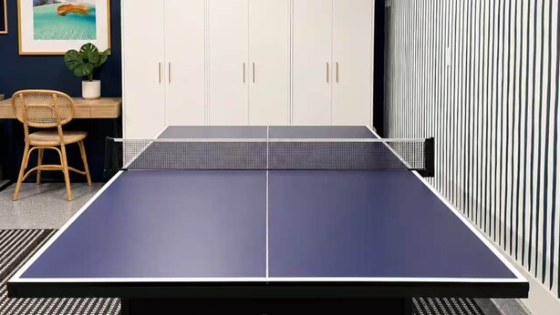 Een kamer met een tafeltennistafel, een wit kastje, een rieten stoel en een lijst met een afbeelding aan de muur.