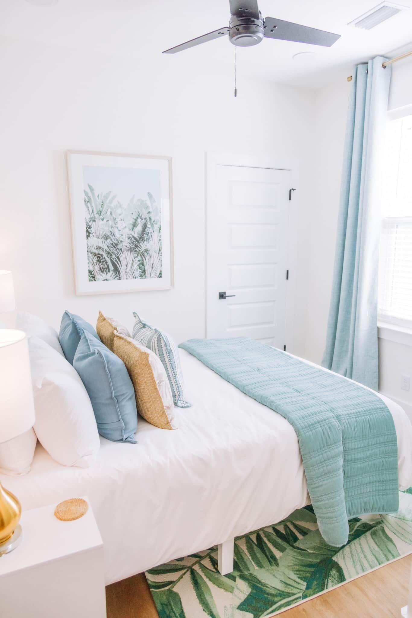 Une chambre à coucher avec un lit, des oreillers, un ventilateur de plafond, une porte et une fenêtre avec des rideaux.