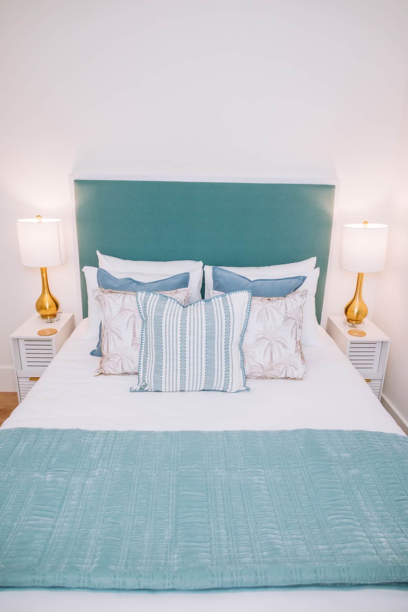 Una cama con cabecera color turquesa, ropa de cama blanca y dos lámparas de noche con bases doradas.