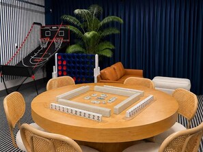 Une table ronde avec des jeux de dominos, un panier de basket et une plante dans une pièce où est accroché un rideau bleu.