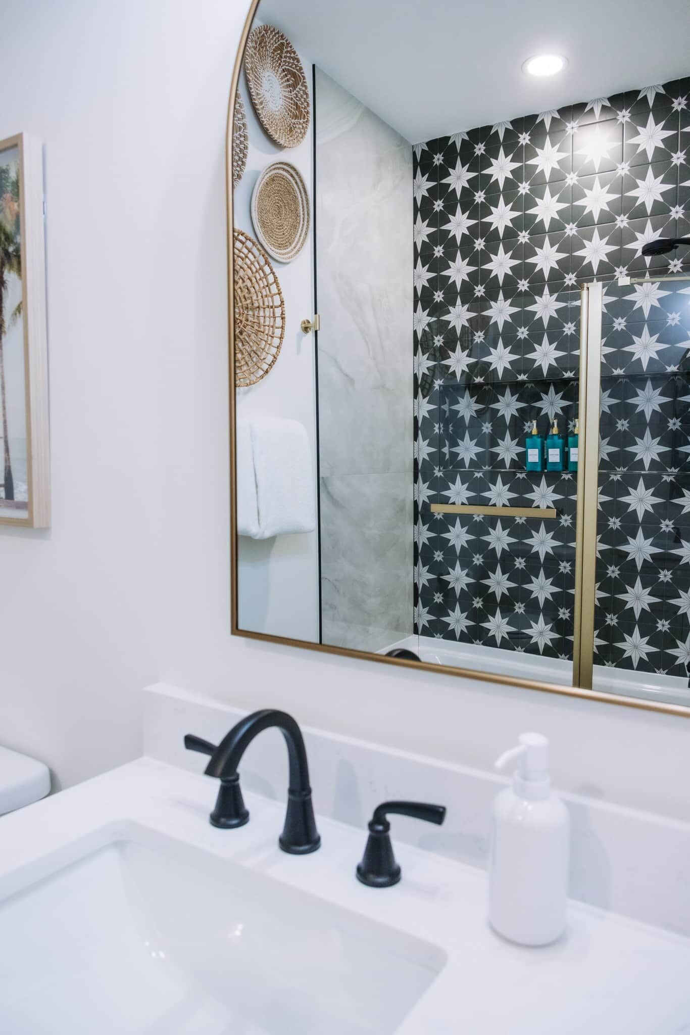 Een badkamer met een wit wastafelmeubel, zwarte kraanwerk, een spiegel en een douche met een betegelde wand in sterrenpatroon.