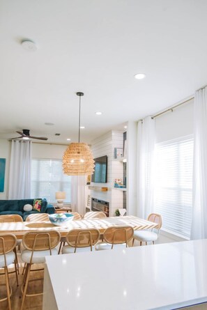 A modern kitchen with white cabinetry, a dining area with a white table and wicker chairs, and a ceiling fan. - The Cove | 4BR · 12 Guests | Spa · Theater · Gym (Walton County)