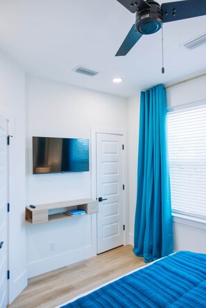 Une chambre à coucher avec un lit, un ventilateur de plafond, un téléviseur fixé au mur, une porte et une fenêtre avec des stores.