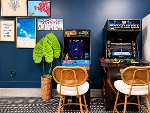 A room with a blue wall, two arcade machines, a large green plant, and two wicker chairs. - The Cove | 4BR · 12 Guests | Spa · Theater · Gym (Walton County)